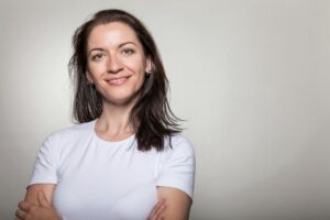 Portrait of a confident woman smiling with arms crossed, wearing a white t-shirt on a gray background.