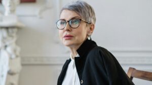 Senior woman with glasses sitting in a stylish office setting, holding a notebook and pen, signifying professionalism.